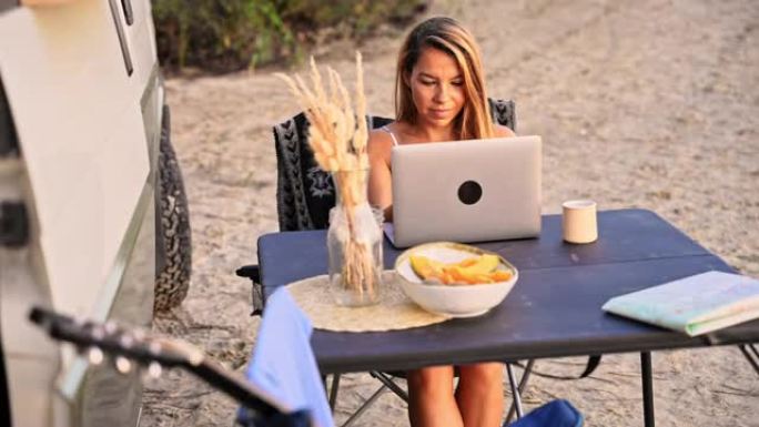 在海滩上日落时，黑发女性在笔记本电脑上工作，旁边是一辆白色野营车