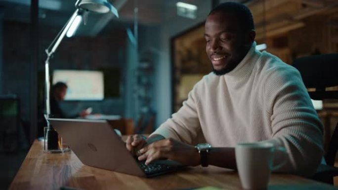 英俊的黑人非洲裔美国男子在创意办公环境中的笔记本电脑上进行在线对话。快乐男性正在浏览社交媒体，并在M