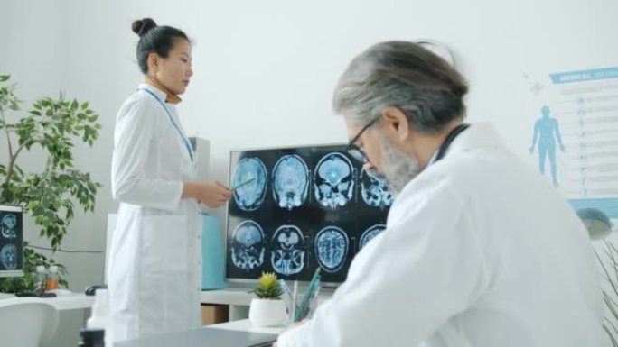 穿着医疗制服的年轻亚洲妇女指着MRI图像并在诊所室内与男性同事交谈