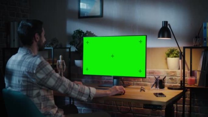 年轻英俊的男人在家工作，在带有绿屏模拟显示的台式计算机上。创意男性查看社交媒体，浏览互联网。晚上阁楼
