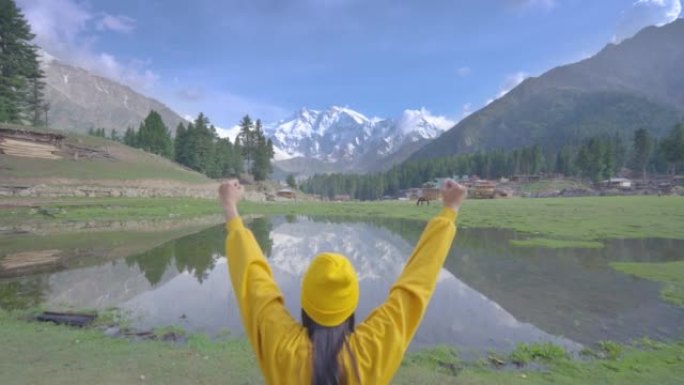 自由快乐的旅行者年轻妇女穿着黄色的休闲布，戴着帽子坐着，举起双臂，一边张开双臂，一边欣赏山上的风景，