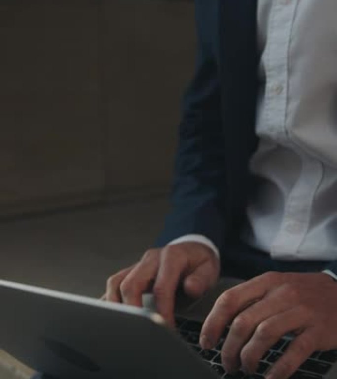 一个穿着蓝布西装和白衬衫的男人的垂直视频，在笔记本电脑上打字，躺在他的腿上，他在街上，只有他的身体和