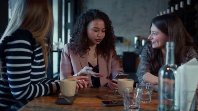友谊概念。三个年轻快乐美丽的多民族女性朋友在时髦的餐厅餐桌上随意交谈。