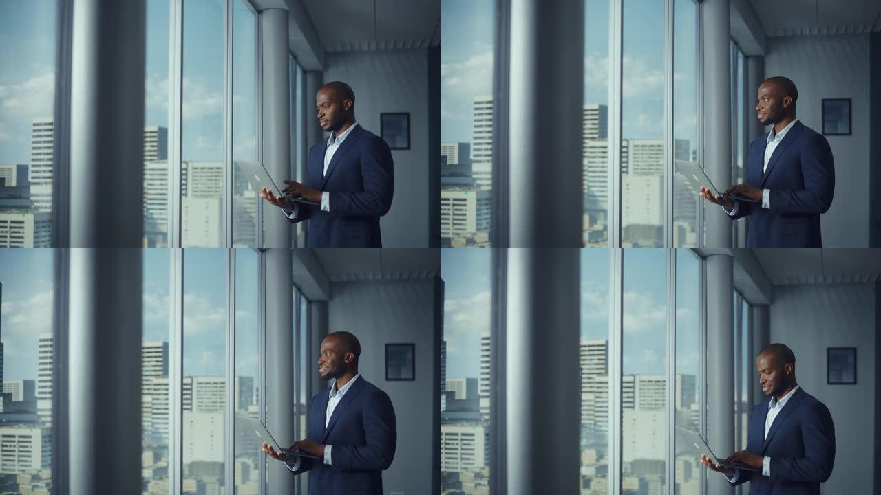 体贴的黑人商人穿着定制的西装，站在大城市窗户附近的办公室里，使用笔记本电脑。成功的企业CEO为电子商