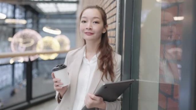 成功的美丽亚洲女商人的肖像，微笑着看着站在办公楼附近的相机，手里拿着笔记本电脑拿着一杯热饮。业务外场