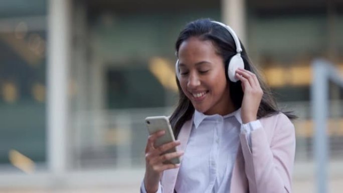 时髦的女人独自一人在城市漫步时使用电话并在耳机上听音乐。酷，时尚，时尚的学生从播放列表中选择和选择城