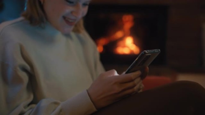 SLO MO DS年轻女子在壁炉前的扶手椅上使用智能手机时微笑