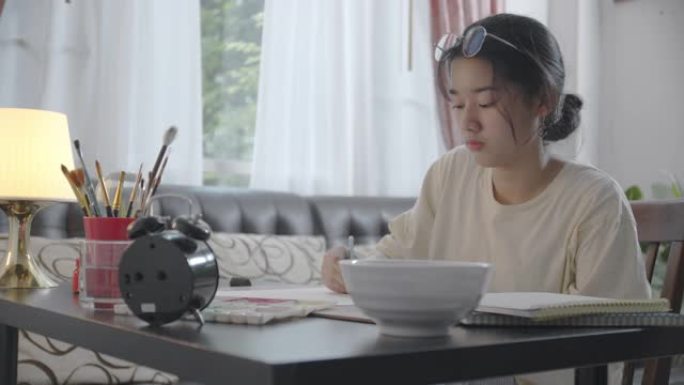 亚洲少女使用笔记本电脑进行视频通话，在家与朋友交谈和微笑，同时画水彩画。她在vlog社交媒体上使用视