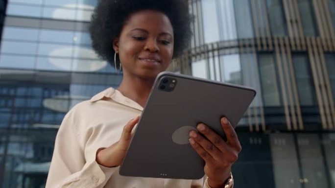 一个年轻的黑皮肤女人，戴着耳环在平板电脑上打字，微笑着，站在现代摩天大楼上，低角度，高腰