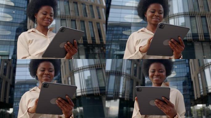 一个年轻的黑皮肤女人，戴着耳环在平板电脑上打字，微笑着，站在现代摩天大楼上，低角度，高腰