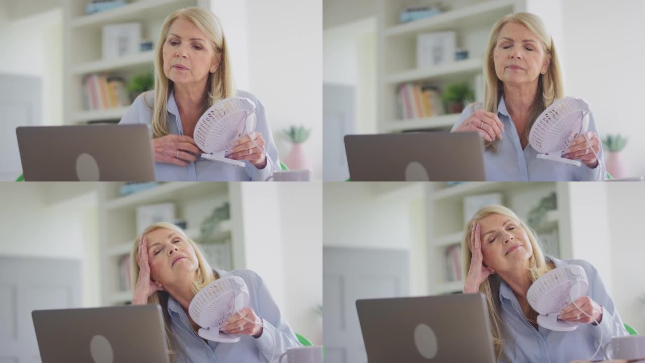更年期成熟的女人在笔记本电脑上工作，家里有连接的风扇，有热冲洗和冷却自己-慢动作拍摄