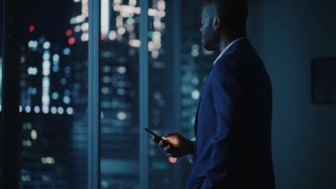 夜间办公室: 成功的黑人商人穿着西装站着，使用智能手机从大城市的窗外望去。非裔美国CEO对电子商务软