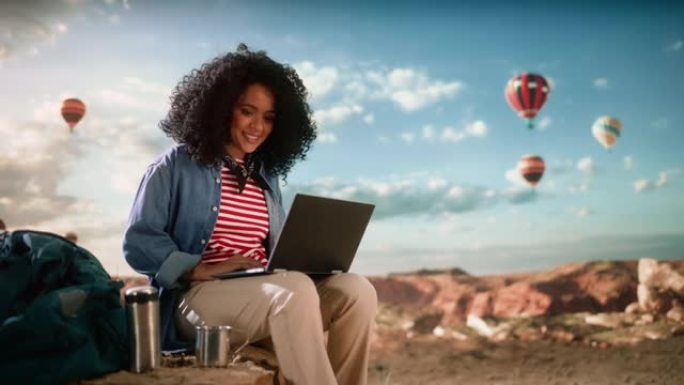在落基峡谷山谷顶部使用笔记本电脑，兴奋的美丽的年轻多民族女性旅行者，拥有非洲发型。热气球在山区国家公