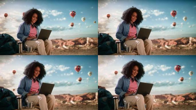 在落基峡谷山谷顶部使用笔记本电脑，兴奋的美丽的年轻多民族女性旅行者，拥有非洲发型。热气球在山区国家公