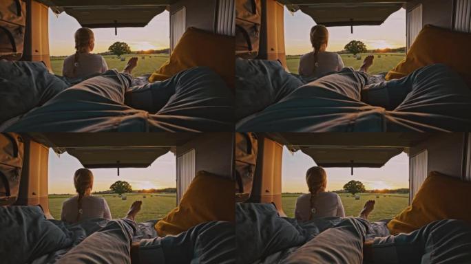 在阳光明媚的日子里，一个男人躺在机舱里，女人坐在停在乡下某个地方的露营者前面