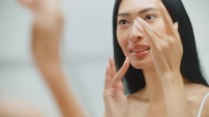 美丽的亚洲女人她完美柔软的面部皮肤，使用先进的有机面霜，微笑在镜子里。快乐的女性享受她早上的例行公事