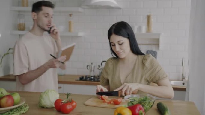 年轻女子正在准备沙拉，而丈夫在手机上讲话并在家里的厨房里吃蔬菜