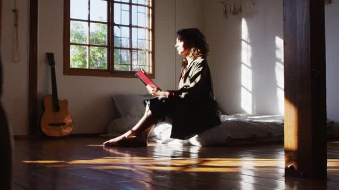 放松的混血女人坐在床上在阳光明媚的小屋卧室看书