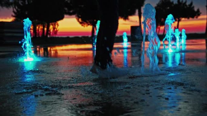 嬉戏的人在城市公园的地面喷泉上享受生活的自由在夏夜户外工作。快乐的男性在日落时玩得开心，在水底跳跃。