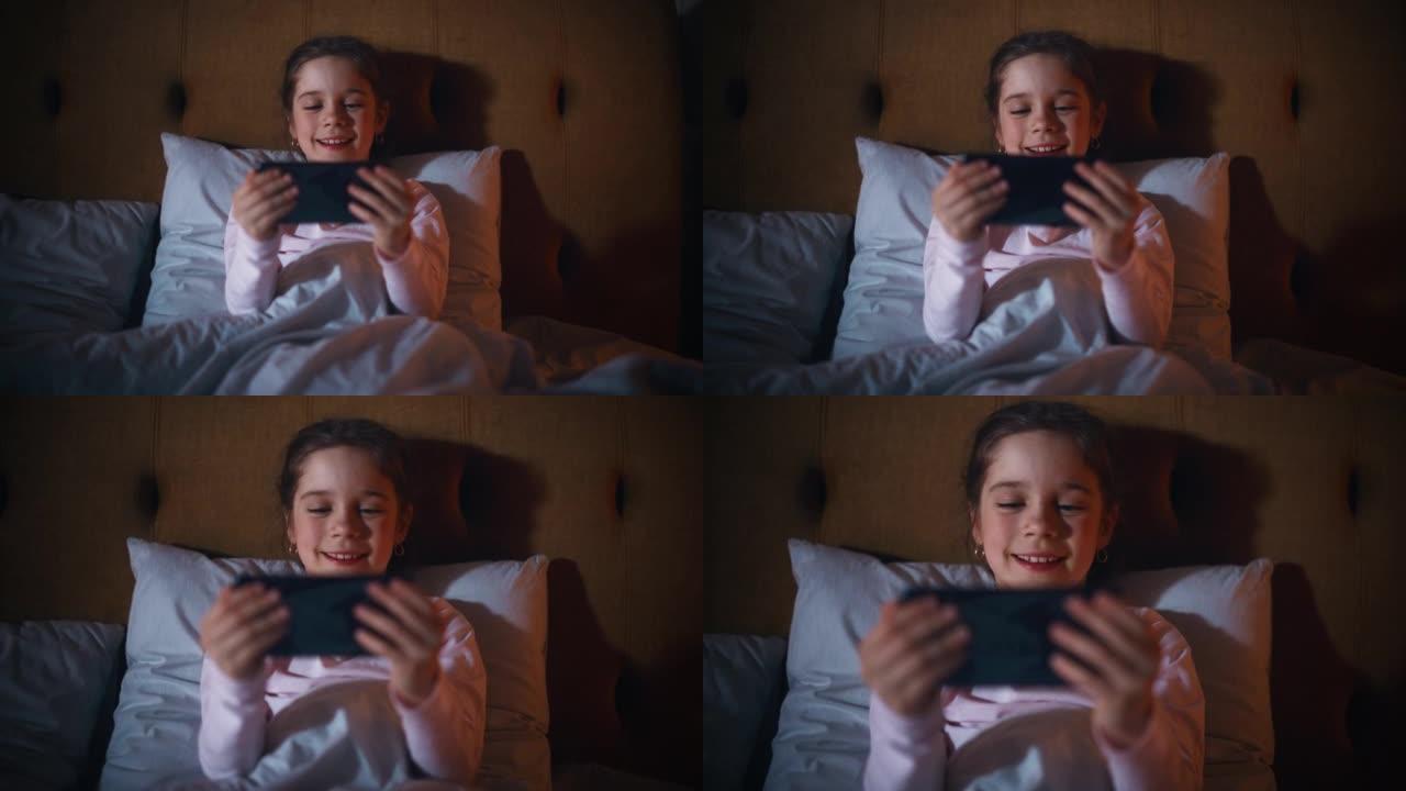 小快乐的女孩睡觉前在智能手机上看娱乐视频。可爱的孩子在社交媒体网站上在线观看卡通和有趣的视频时笑。