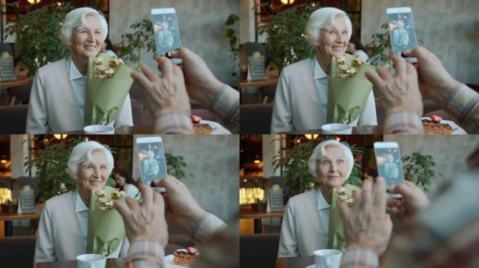 美丽的退休妇女在餐厅里为智能手机相机摆姿势，拿着鲜花