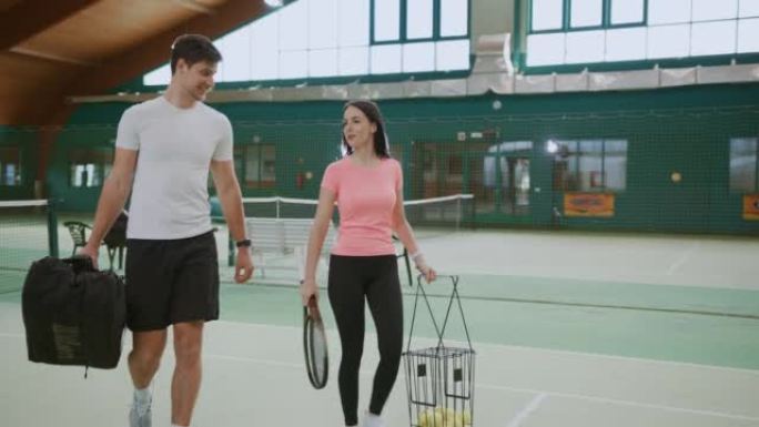 年轻的男教练和带网球设备的妇女走进室内网球场