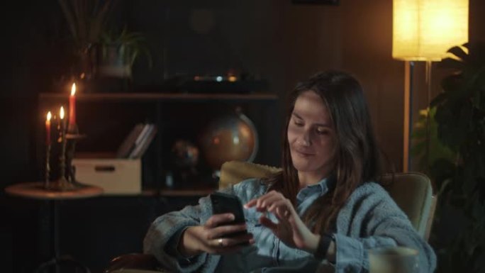 使用智能手机社交媒体应用程序在家喝茶的美丽年轻快乐微笑女人的电影黑暗肖像。