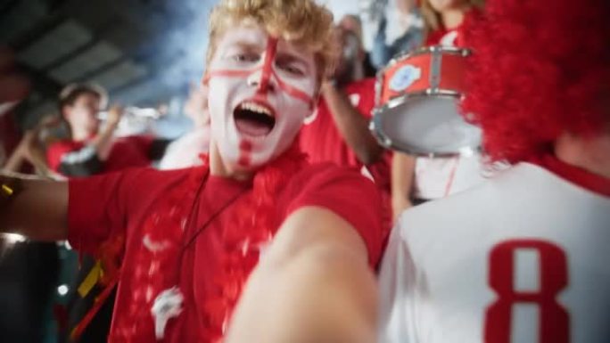 体育场体育赛事: 画脸男子的自拍肖像拿着智能手机开枪，为红队获胜欢呼，尖叫，与人群一起庆祝冠军胜利