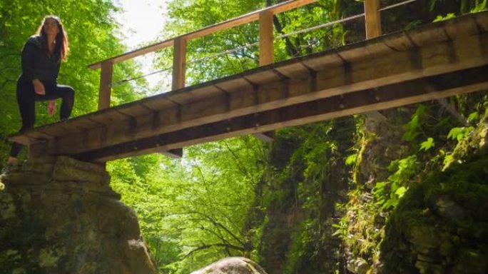 在阳光明媚的日子里，有身体意识的女人在绿色景观的山溪峡谷上的木桥上徒步旅行