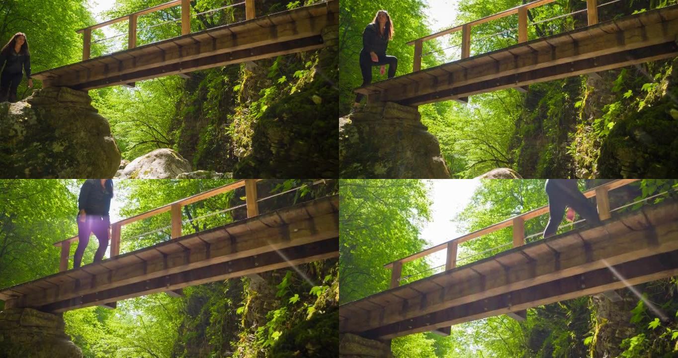 在阳光明媚的日子里，有身体意识的女人在绿色景观的山溪峡谷上的木桥上徒步旅行