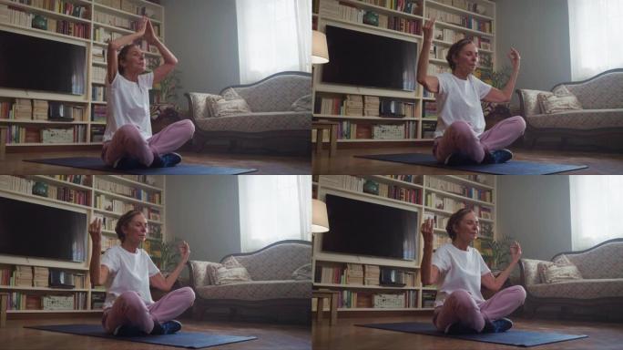 电影拍摄的宁静微笑的成熟女性正在垫子上练习瑜伽，并在家里的客厅放松自己的思想。健康生活方式、精神平衡