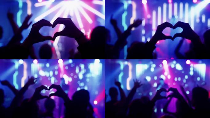 人正在做一个心形手势，并在表演中举手。摇滚乐队在舞台上的夜总会音乐会上演奏一首歌，灯光鲜艳。