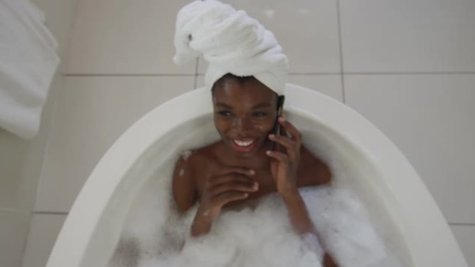 微笑的非洲裔美国迷人的女人在浴室放松，在浴室用智能手机聊天