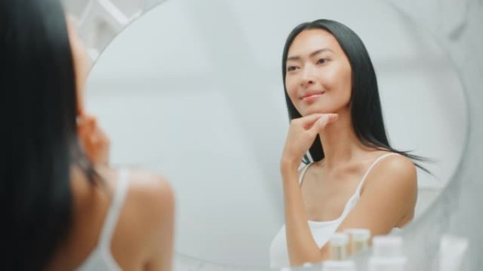美丽的亚洲女人扮演和欣赏她完美柔软的皮肤和郁郁葱葱的黑发，看着相机，在浴室的镜子里微笑。健康概念，自