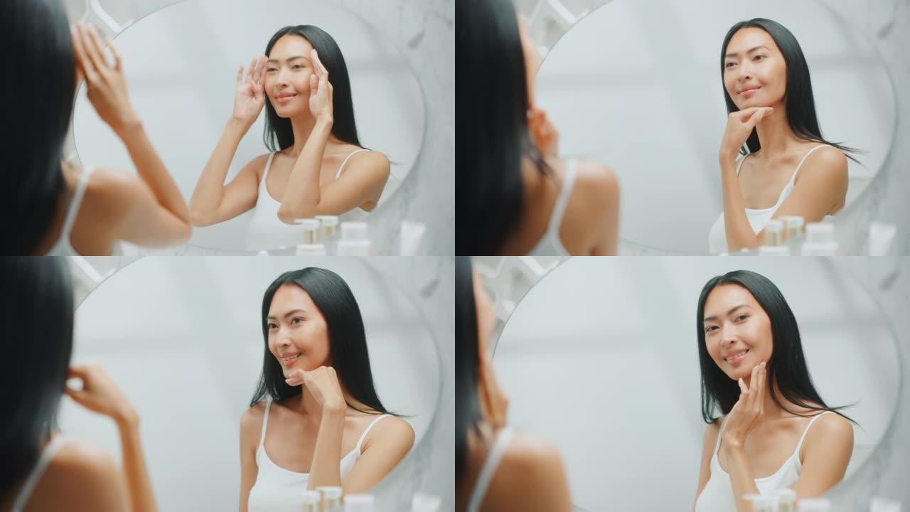 美丽的亚洲女人扮演和欣赏她完美柔软的皮肤和郁郁葱葱的黑发，看着相机，在浴室的镜子里微笑。健康概念，自