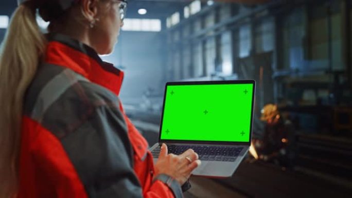 专业重工业工程师使用带有绿屏模拟显示的笔记本电脑。在金属制造仓库工作的女性工业专家，背景是火花。