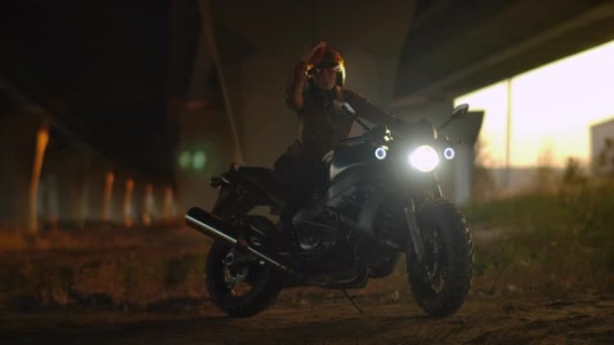 穿着皮夹克的骑自行车的女人戴着摩托车镀铬头盔。性感的司机女孩戴着头盔坐在城市的摩托车上。美丽的女性微