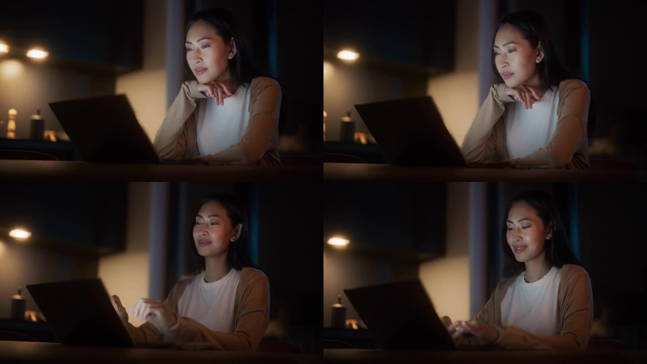 美丽地道的亚洲女人坐在舒适的厨房里的桌子旁，晚上在家使用笔记本电脑。女性在社交媒体上微笑聊天，网上购