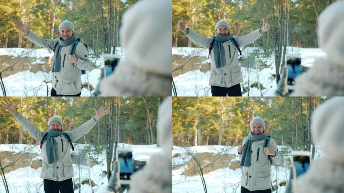 快乐的老人游客在冬季在森林中摆出智能手机相机的姿势，显示竖起大拇指的手势