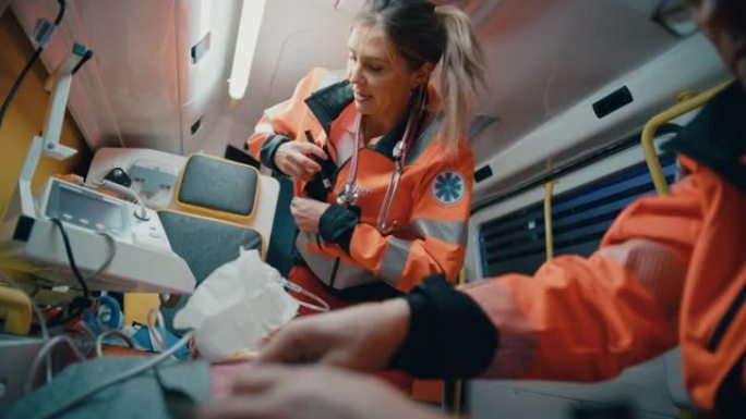 女性和男性EMS护理人员提供医疗帮助受伤的病人在去医疗保健医院的路上。急救助理在救护车中用手电筒检查