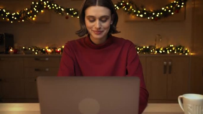 一位穿着红色毛衣的迷人年轻女子坐在圣诞节装饰的厨房的桌子旁，在笔记本电脑上打字