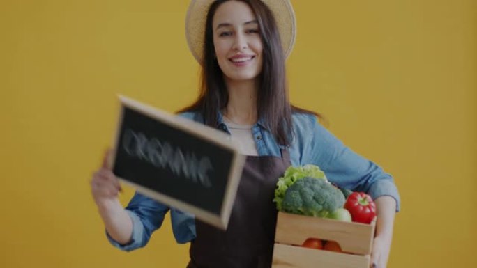 年轻女子拿着盒子的肖像，黄色背景上有新鲜蔬菜举起标志有机
