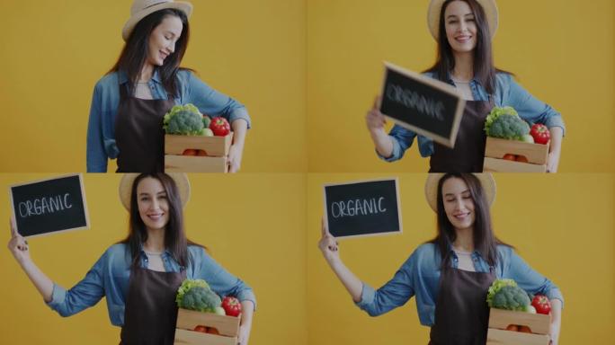 年轻女子拿着盒子的肖像，黄色背景上有新鲜蔬菜举起标志有机
