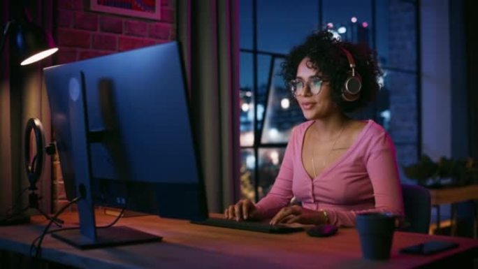 年轻迷人的黑人妇女晚上在时尚的阁楼公寓里使用电脑。富有创造力的女性微笑，查看社交媒体，浏览互联网上的