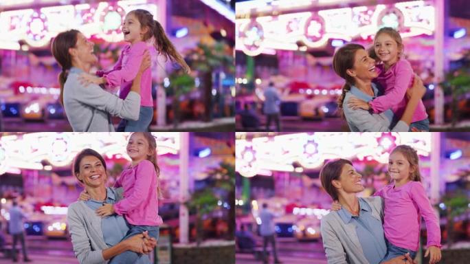 一个快乐的母亲和她的小女儿的真实照片在一起玩得开心，晚上在游乐园里带着露娜公园的灯光在相机里微笑。