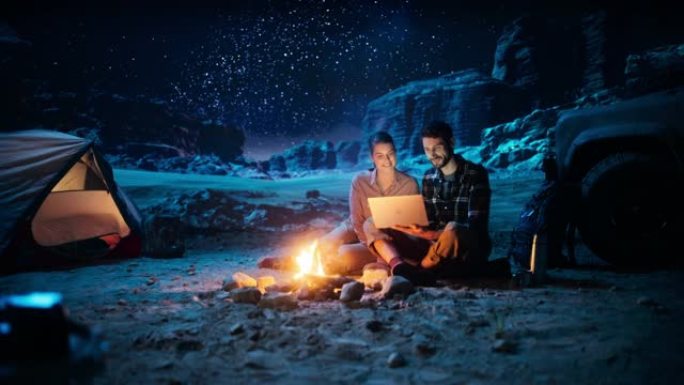 晚上在峡谷露营的快乐夫妇，坐在篝火旁使用笔记本电脑。两个人在社交媒体上发表鼓舞人心的文章，使用互联网