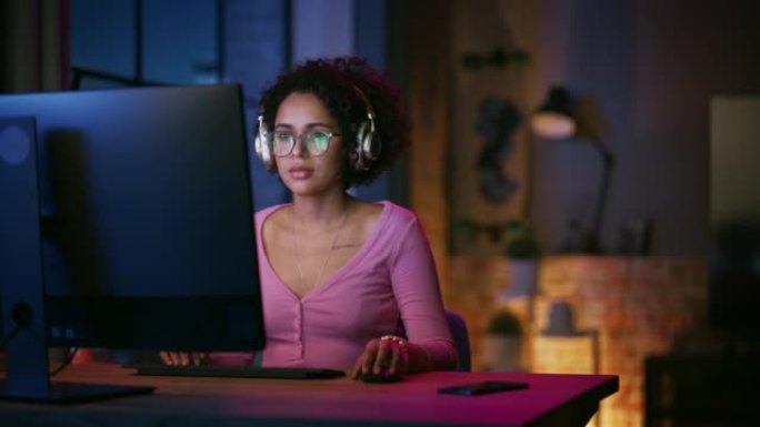 年轻迷人的黑人女性晚上在时尚的阁楼公寓里使用台式电脑。富有创造力的女性微笑，查看社交媒体，浏览互联网