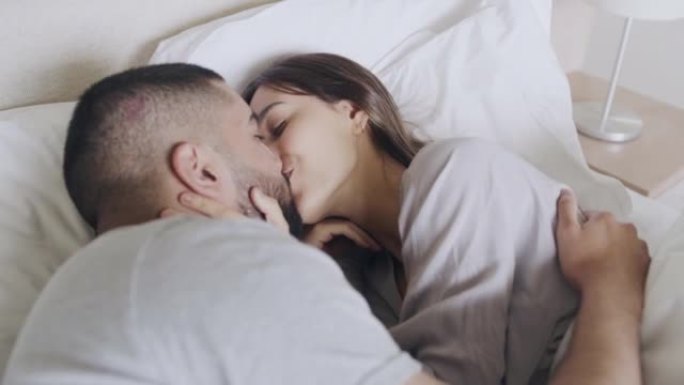 亲爱的夫妇躺在床上互相亲吻早上好。幸福的夫妻在现代卧室里拥抱在床上。年轻的已婚夫妇在新家一起醒来