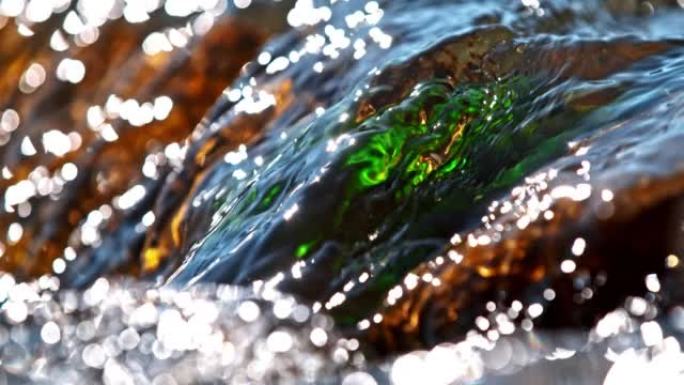超级SLO MO特写水从河中的棕色绿色大石头流下，水面上的阳光反射