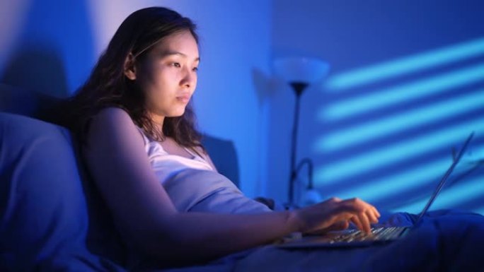 年轻的亚洲妇女工作到很晚，在床上使用笔记本电脑